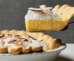  A slice of coconut cream pie shown from the side on a pie lifter, freshly cut from a full pie in a decorative white baking dish, topped with whipped cream and shaved coconut.