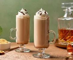 Two tall glasses of Irish whiskey latte topped with whipped cream and chocolate shavings, presented on a wooden table with a side of brown sugar, a decanter of amber whiskey in the background.