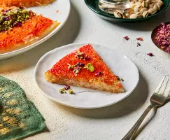 A slice of knafeh on a plate, garnished with pistachios and edible flower petals, shown with a full knafeh, dishes of pistachios in the shell and flower petals and a lit candle. 