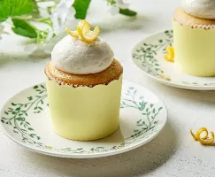 Lemon cupcakes with a fluffy vanilla frosting, adorned with a twist of lemon zest, presented on a vintage floral-patterned plate set against a white wooden surface and a backdrop of soft greenery and white blooms for a fresh, springtime feel.