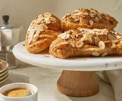 Four air fryer almond croissants topped with sliced almonds on a white cake stand, with an espresso cup and stovetop coffee maker in the background.