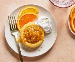 Overhead shot of a single sponge cake topped with glistening citrus marmalade and zest, served on a white ridged plate with fresh orange slices and a dollop of whipped cream, accompanied by a fork, against a soft peach-toned backdrop.