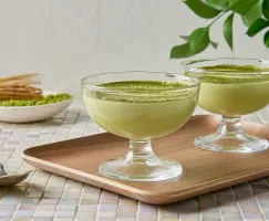  Two glass cups of white chocolate matcha mousse garnished with green matcha powder on a wooden tray, with a traditional matcha whisk and bowl of matcha powder in the background, set on a tiled table with natural light.