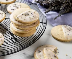 A batch of glazed lemon-lavender shortbread cookies sprinkled with dried lavender, shown on a circular wire cooling rack on a marble counter, with dried lavender stalks and a lavender-coloured cloth.