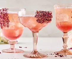 Three unique stemmed glasses of lychee-rose spritzer on ice garnished with dried rose petals on the glasses, shown in a neutral setting with fresh roses in the background and dried rose petals scattered around.