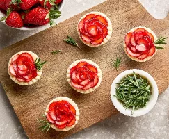 Five strawberry rosemary tartlets on a wooden cutting board, shown with a bowl of rosemary sprigs, a bowl of fresh strawberries, and a sixth tart on the counter.