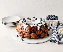 Blueberry monkey bread on a white plate with dripping white icing and fresh blueberries on top, next to a blue and white striped napkin, blue bowl with icing, and a carton of blueberries, on a light-coloured surface.