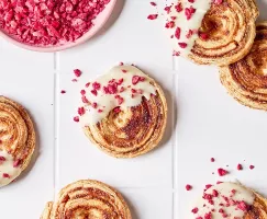 Overhead view of raspberry puff pastry cookies with white chocolate icing and freeze-dried raspberry pieces on a white tiled countertop, shown with a bowl of crushed raspberries.