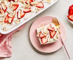 A single serving of Strawberry-Coconut Tres Leches cake on a pink plate with a gold fork, alongside a white baking dish of the same cake and a small bowl of sliced strawberries, all on a grey countertop with a pink cloth napkin