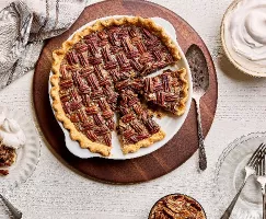 A pecan pie in a baking dish, shown with three slices cut, with one served on a plate with whipped cream, on a white wood table with a bowl of whipped cream and a bowl of pecans.