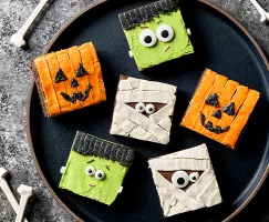 Six square brownies decorated for Halloween like jack o’lanterns, mummies, and Frankenstein, shown on a black plate with bones scattered around.