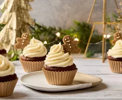 Festive Gingerbread Cupcakes topped with a swirl of creamy white frosting and a mini gingerbread cookie. The cupcakes are arranged on a wooden surface with Christmas decorations in the background, including a gold Christmas tree and twinkling fairy lights.