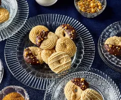 Assortment of piped butter cookies presented on glass plates, showcasing three decoration styles: half-dipped in chocolate with gold sprinkles, drizzled with white chocolate, and sprinkled with golden sugar. The background is a dark blue tablecloth, with additional golden sprinkles in a small dish.