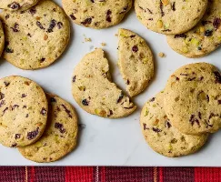 Cranberry pistachio cookies arranged on a white marble surface. The slice-and-bake cookies feature visible chunks of dried cranberries and pistachios, adding texture and colour. One cookie is split in half, showing a tender crumb. A red plaid cloth adds a festive touch at the bottom of the image