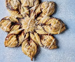 A beautifully crafted Glazed Cinnamon Sugar Snowflake pastry, shaped like a snowflake with twisted layers of dough, filled with cinnamon and sugar. The pastry is drizzled with a light glaze and dusted with powdered sugar, shown on a pale blue background.