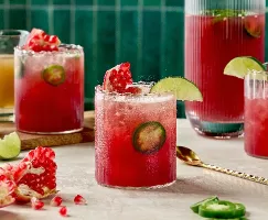 Three glasses of Spicy Pomegranate Ginger Fizz, featuring a vibrant red hue with slices of fresh jalapeño. The drinks are garnished with lime wedges and pomegranate seeds. A pitcher filled with more of the drink stands in the background, alongside lime wedges, pomegranate pieces, and a small pitcher of ginger syrup. The scene is set against an emerald green-tiled backdrop.