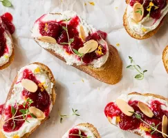 A close-up view of several crostini topped with whipped feta cheese, cranberry chutney, slivered almonds, and fresh thyme.