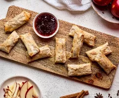 A wooden cutting board holds a pile of golden brown egg rolls. A small bowl of strawberry jam sits nearby. Sliced apples and cinnamon sticks are scattered around the board, suggesting the egg rolls are filled with apple and cinnamon.