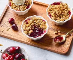 Individual servings of Cherry Buckwheat Crisp in white ramekins, topped with golden oat crumble, one with a scoop of vanilla ice cream. Fresh cherries are scattered on the wooden serving tray, with a bowl of cherries nearby and a gold spoon holding some of the juicy filling, highlighting the dessert's vibrant color and texture.
