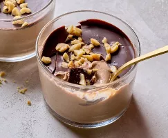  Close-up of creamy chocolate peanut butter pudding in a glass jar, topped with a glossy chocolate layer and chopped peanuts. A golden spoon rests inside, revealing the smooth and indulgent texture beneath the chocolate shell. Crumbled peanuts are scattered on the light gray surface.
