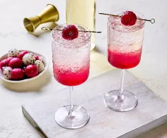 Two glasses of vibrant dragon fruit spritzer served over ice, with a pink gradient from the fruit at the bottom. Each glass is garnished with a skewered dragon fruit ball. A plate of red and white dragon fruit balls, a gold jigger, and a bottle are visible in the background.