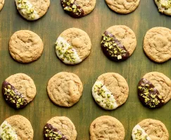 Chocolate chipless cookies arranged in a grid pattern on a green wooden surface. Some cookies are plain, while others are half-dipped in white or dark chocolate and sprinkled with chopped pistachios. 