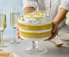 A person’s hands hold a glass trifle dish filled with a layered lemon elderflower trifle featuring alternating layers of sponge cake, lemon curd, and whipped cream. The trifle is topped with lemon zest curls and delicate white flowers. In the background are champagne glasses, a bottle, and white roses in a glass vase.