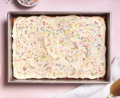 A rectangular frosted cake covered in colorful sprinkles sits in a metal baking pan on a light pink surface, with a small bowl of more sprinkles, plates and forks, and a gold cake server nearby.