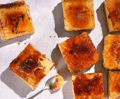 An overhead shot of several small, square slices of caramelized cheesecake with a burnt sugar topping, scattered on a white surface with a spoon taking a bite out of one piece.