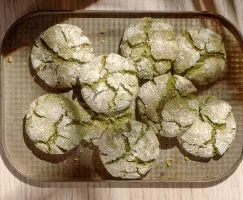 Eight pale green matcha crinkle cookies, coated in powdered sugar with characteristic cracks, arranged on a rectangular metal baking sheet.