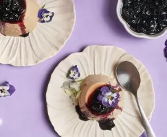  Top-down view of two servings of Earl Grey panna cotta topped with blueberry compote and decorated with purple and white edible flowers, served on scalloped cream plates against a lavender background with scattered blossoms and a spoon.