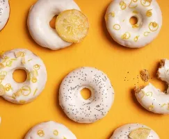 Glazed lemon poppyseed doughnuts arranged on a vibrant yellow background, decorated with lemon zest, candied lemon slices, and poppy seeds; one doughnut has a bite taken out.
