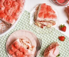 A white baking dish of strawberries and cream tiramisu on a green gingham cloth on a white tiled surface with two slices cut out and served on pink and clear glass plates, surrounded by fresh strawberries and pink roses.