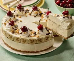 A creamy banana split ice cream pie topped with swirls of whipped cream, cherries, chopped nuts, and chocolate shavings sits on a white plate. A slice is being lifted to reveal layers of banana slices and frozen filling over a graham cracker crust. In the background, there’s a stack of pastel plates with forks and a bowl of fresh cherries on a light green tablecloth.