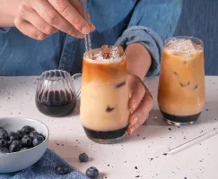 Person in a denim shirt stirring a glass of iced blueberry coffee with a glass straw. The drink has distinct layers of blueberry syrup, milk, and coffee. A second identical drink sits nearby on a speckled countertop. A glass pitcher of blueberry syrup, a bowl of fresh blueberries, and scattered berries complete the scene.