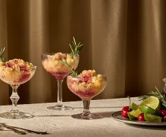 Three coupe glasses filled with a layered frozen dessert of golden brown and ruby red slush, garnished with fresh rosemary sprigs, are set on a light textured surface in front of a warm brown curtain. Beside them sits a plate with fresh limes, lime wedges, cherries, and rosemary, as well as a small glass pitcher of dark cherry syrup