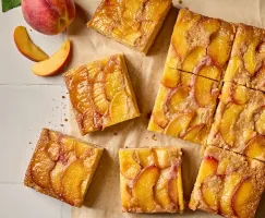 Top-down view of golden-brown peach upside-down cake squares arranged on a sheet of parchment paper. The squares are topped with glistening caramelized peach slices. A whole fresh peach and a few peach wedges are placed decoratively beside the cake pieces.