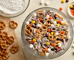 Overhead view of a large glass bowl filled with Puppy Chow, a chocolate-coated cereal and pretzel snack dusted in icing sugar. Surrounding the bowl are scattered pretzels on parchment paper, a bowl of icing sugar, and a small dish of chocolate chips.