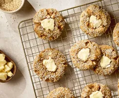 A top-down shot shows several white chocolate tahini cookies with a visible texture from being coated in white sesame seeds, arranged on a brass-colored wire cooling rack. Each cookie has an indent in the center filled with a dollop of melted white chocolate or cream cheese frosting. In the upper left corner, a small bowl holds more white sesame seeds. In the lower left, a small brown dish contains irregular chunks of white chocolate. The background is a light, textured surface