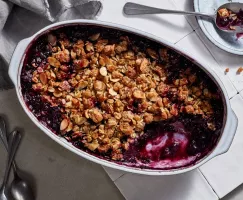 Serving dish full of blueberry crisp with a small amount scooped out on a tile counter