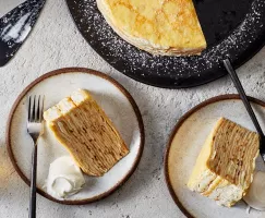 Two slices of bourbon crêpe cake on plates served with whipped cream and a glass of bourbon