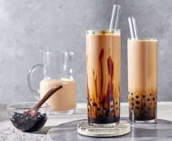  Two glasses of boba hojicha milk tea shown with a pitcher of tea and a bowl of tapioca pearls