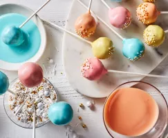 Pastel-coloured cake pops with sprinkles on a platter, some standing in a glass of sprinkles, and one being dipped in blue icing