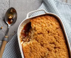 pudding chomeur in a dish with a scoop missing, the dish is sitting on a blue checkered napkin