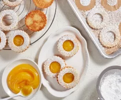 Citrus curd cookies on a platter shown with unfinished cookies on a cooling rack and a baking pan and a bowl of citrus curd