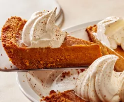 Removing a slice of pumpkin pie with whipped cream and ground cinnamon from a white pie plate with a pie lifter.  