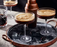 Two glasses of frothy espresso martini on a tray with a cocktail shaker with a third glass and a glass pitcher of espresso marti