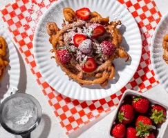 Funnel cake dusted with icing sugar and topped with strawberries on a paper plate