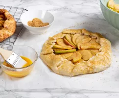 Unbaked apple galette sprinkled with turbinado sugar, and egg wash with a brush in a bowl