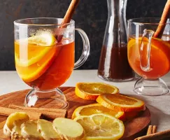 Two hot toddies in glass mugs garnished with orange slices and cinnamon sticks, one on a wood platter with garnishes and sliced